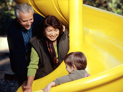 Hispanic older adult grandparents watching grandson go down a yellow slide.
